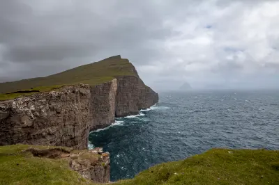 Vertical sea cliffs at Trælanípa on Vágar Island in the Faroe Islands, with basalt rock walls rising above rough Atlantic waters and Mykines Island visible on the horizon.
