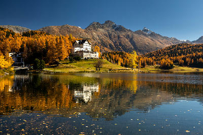 Lake St. Moritz with autumn larch forest, lakeside hotel, and alpine peaks reflected in calm water, Engadin, Switzerland.
