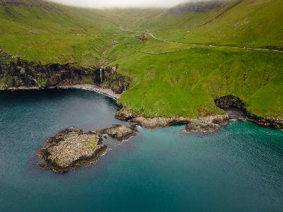 Wide aerial view of Skarðsáfossur waterfall descending over basalt cliffs into a sheltered Atlantic cove on Vágar Island, Faroe Islands.
