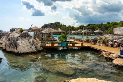 Rocky waterfront in Kaleköy near Demre, Turkey, featuring wooden platforms, straw parasols and sun loungers beside calm, transparent turquoise water under a dramatic summer sky.