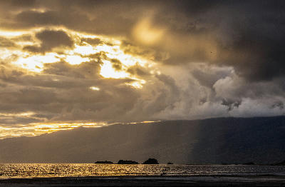 Golden sunlight breaking through dramatic storm clouds above Lake Natron with rocky islets and the Rift Valley escarpment in northern Tanzania.
