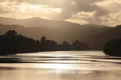 Afternoon sunlight breaks through cloudy skies, casting a bright golden shimmer across the wide Daintree River as silhouettes of forested hills rise in the background.