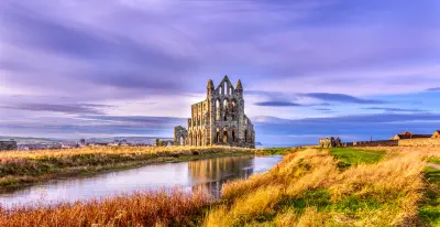 Wide view of Whitby Abbey's Gothic ruins beneath a purple-blue evening sky, reflected in a pool amid golden grasses on the East Cliff, North Yorkshire, England.