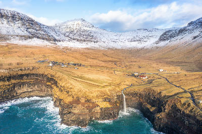 Aerial view of Gásadalur village and Múlafossur Waterfall dropping into the Atlantic beneath snow-dusted mountains on Vágar Island, Faroe Islands.
