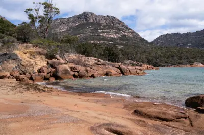 Sandy shoreline, clear water and pink-orange granite rocks at Honeymoon Bay with a rugged peak of The Hazards rising behind, Freycinet National Park, Tasmania.