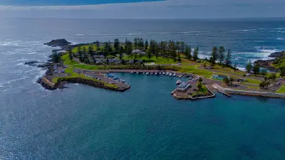 Aerial view across Kiama Harbour toward the pine-lined Blowhole Point, surrounded by rocky coastline and the blue Pacific Ocean.