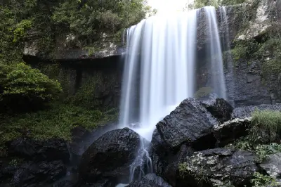 Frontal view of Zillie Falls cascading over a broad rocky cliff into a gorge filled with large dark boulders and lush rainforest in Queensland, Australia.