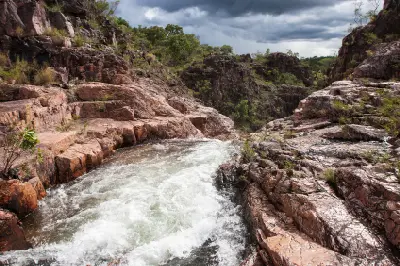 White water rushing through a narrow sandstone channel above Tolmer Falls, surrounded by rugged reddish rocks and sparse woodland beneath dark storm clouds in Litchfield National Park, Northern Territory, Australia.
