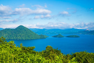 Elevated view of Lake Toya with forested islands in the blue water and surrounding mountains under a partly cloudy sky.