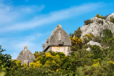 Ancient Lycian stone sarcophagi surrounded by green shrubs and yellow wildflowers on a rocky hillside in Simena, Kaleköy, Kekova, Antalya, Turkey.