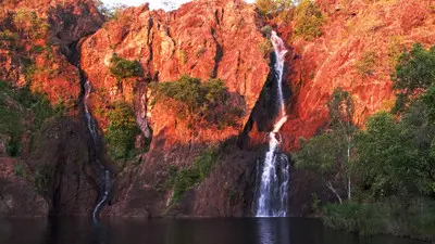 Two waterfalls cascading down vivid red sandstone cliffs into a dark plunge pool at sunset at Wangi Falls, Litchfield National Park, Northern Territory, Australia.
