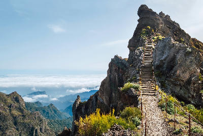 Stone steps climbing a narrow volcanic ridge along the Pico do Arieiro–Pico Ruivo route near Pico do Arieiro, with steep mountain slopes and distant cloud layers below.
