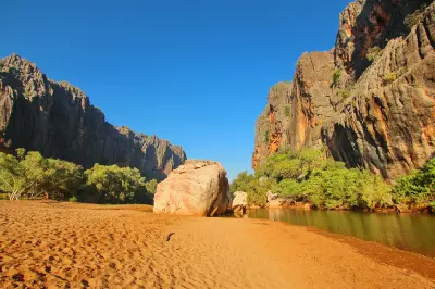 Sandy riverbank and calm water beneath towering dark limestone cliffs and orange rock faces in Bandilngan (Windjana Gorge).