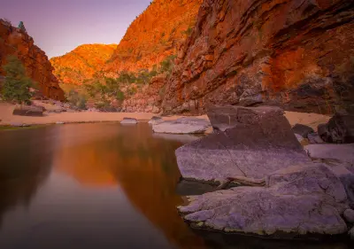 Calm waterhole and large foreground rocks beneath towering red cliffs glowing in warm light at Ormiston Gorge.