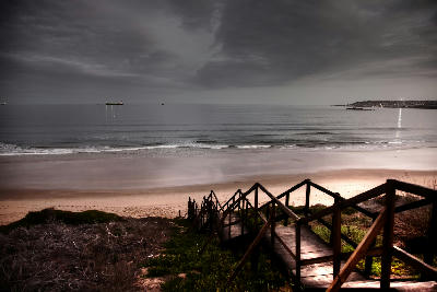 Boardwalk steps leading down to Santos Beach at dusk with calm ocean waters and ships anchored offshore in Mossel Bay, South Africa.