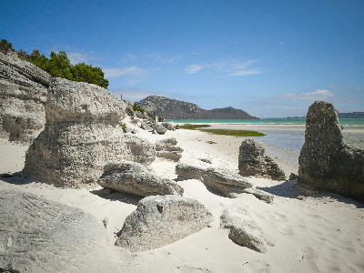 White sandstone formations and soft sand along the shoreline of the Langebaan Lagoon in West Coast National Park, with turquoise water and low coastal hills in the background.