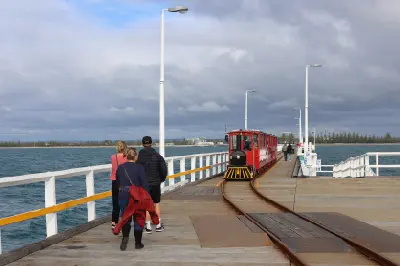 Red tourist train travelling along Busselton Jetty with visitors walking beside the railway tracks.