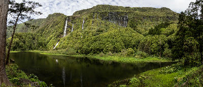 Panoramic view of Poço da Ribeira do Ferreiro on Flores Island, with multiple waterfalls descending lush green cliffs into a calm reflective lagoon in the Azores.