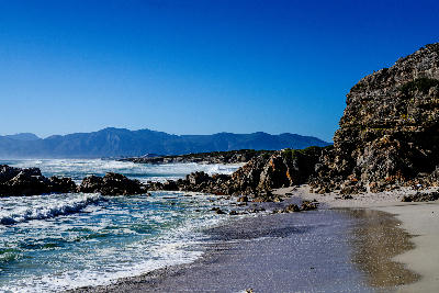 Surf breaking along the rocky De Kelders coastline with cliffs, sandy shoreline, and distant mountains beside Walker Bay, South Africa.