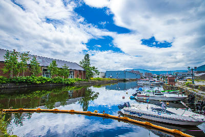 Boats moored along Otaru Canal with warehouse buildings and reflections under a partly cloudy sky in Hokkaido, Japan.