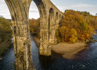 Close aerial view of Lambley Viaduct's massive stone piers and repeating arches crossing the River South Tyne amid colourful autumn trees.
