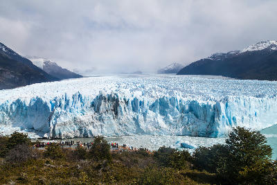 Wide view of Perito Moreno Glacier ice wall with tourists on boardwalks and mountains in the background in Patagonia, Argentina.