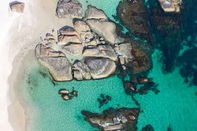 Bird's-eye view of massive weathered rock formations resting in shallow, clear turquoise sea along a sandy shoreline.