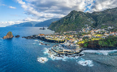 Aerial view of Porto Moniz village on Madeira Island, showing volcanic lava pools, black basalt coastline, and steep green cliffs above the Atlantic Ocean.