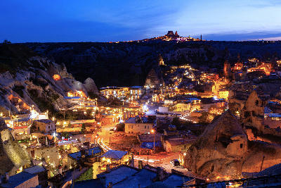 Night view of Göreme in Cappadocia, Turkey, with illuminated cave hotels, fairy chimneys, and rock-cut buildings beneath a deep blue twilight sky, with Uçhisar Castle visible in the distance.