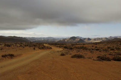 A remote dirt road crosses the desert plains of the Richtersveld, with rugged mountains stretching across the South Africa–Namibia border beneath dramatic storm clouds.