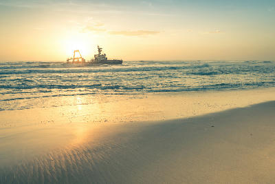 Sunset view of the Zeila shipwreck off Namibia’s Skeleton Coast with waves rolling onto a sandy beach in the foreground.