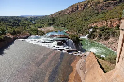 View from Hartbeespoort Dam showing water flowing over the spillway into the Crocodile River, with waterfalls, rocky cliffs and dense green vegetation in the Magaliesberg valley.