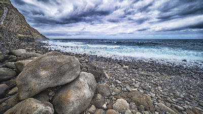 Rocky coastline at Calhau de São Jorge near Santana, Madeira, with large boulders, pebble beach, and Atlantic waves under dark, stormy clouds.
