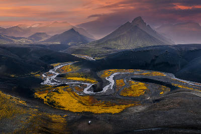 Aerial view of a braided glacial river flowing through black sand and yellow moss near Landmannalaugar in the Iceland Highlands, with mountains and pink-orange evening clouds in the background.