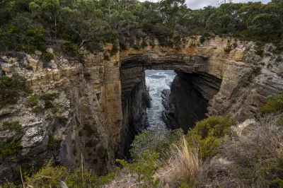 View from above of Tasman Arch spanning a narrow ocean channel between layered cliffs on the Tasman Peninsula, Tasmania.