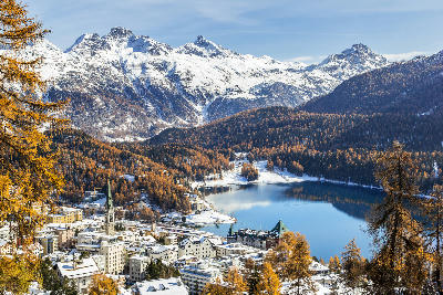 Elevated view of St. Moritz and Lake St. Moritz with autumn larch trees, light snow cover, and surrounding alpine mountains in the Upper Engadin, Switzerland.