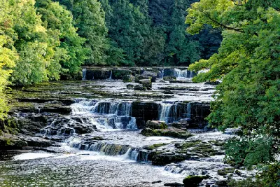 Multiple stepped limestone waterfalls along the River Ure at Aysgarth Upper Falls, framed by green woodland in the Yorkshire Dales National Park.
