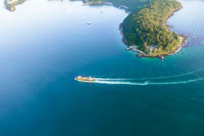 Aerial view of Bradleys Head extending into Sydney Harbour, with a ferry curving through the blue water past the forested peninsula.