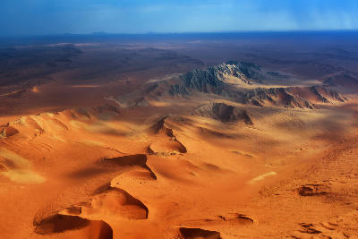 High aerial view of sweeping orange-red sand dunes and rugged desert mountains in the Namib Desert under dramatic light in Namibia.
