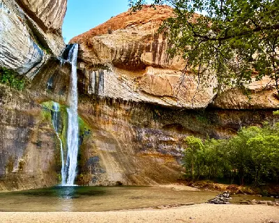 Lower Calf Creek Falls drops over a sweeping sandstone cliff into a clear pool surrounded by green vegetation in the Utah desert.