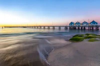 Busselton Jetty extending across calm water at sunset, with its blue buildings and a pastel sky over Geographe Bay.