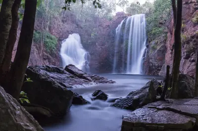 Atmospheric early morning view of the twin waterfalls at Florence Falls, framed by dark rocks and tropical trees, with silky flowing water entering the plunge pool in Litchfield National Park, Northern Territory, Australia.