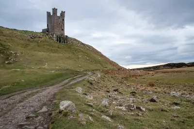 A rocky walking path winding towards a ruined tower of Dunstanburgh Castle, with grassy slopes and an overcast sky on the Northumberland coast.