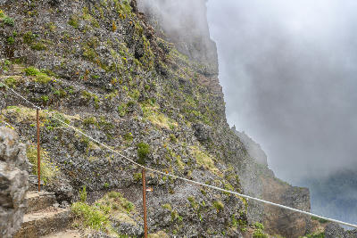 Narrow mountain path carved into volcanic rock near Pico do Arieiro, with safety cables and clouds drifting across steep cliffs along the island’s central ridge.