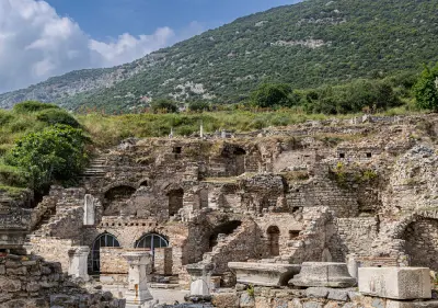 Stone and brick ruins of the Terrace Houses beside Curetes Street in ancient Ephesus, with a green mountainside rising behind.