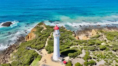 Aerial view of Split Point Lighthouse with its red cap rising above coastal heath and turquoise ocean at Aireys Inlet on Victoria’s Great Ocean Road.