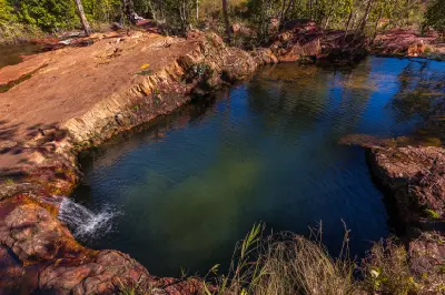 Elevated view of a deep blue-green natural rock pool bordered by reddish sandstone, with a small cascade flowing into the water at Buley Rockhole, Northern Territory, Australia.
