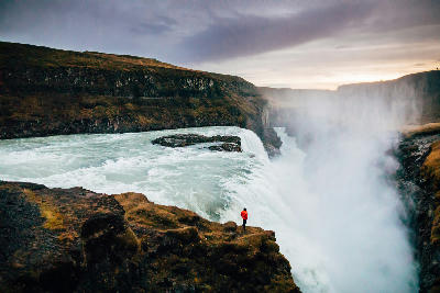 Gullfoss waterfall on the Golden Circle in Iceland, showing powerful white water cascading into a mist-filled canyon with a person standing on the cliff for scale.