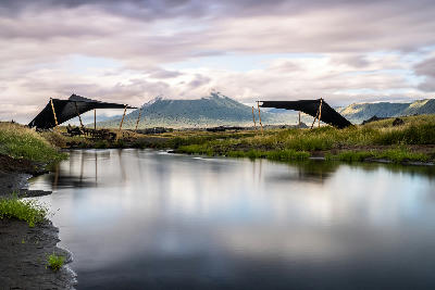 Safari camp beside a reflective stream with Ol Doinyo Lengai rising in the background under low clouds near Lake Natron in northern Tanzania.