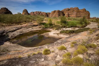 Striped sandstone domes rise beyond a shallow waterhole and spinifex vegetation near Cathedral Gorge in the Bungle Bungles.